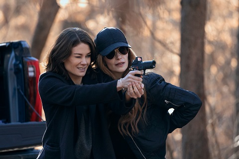 "Two women in dark clothing stand outdoors in a wooded area; one is teaching or assisting the other in aiming a handgun. A pickup truck bed is visible behind them, and the background features blurred trees in daylight."
