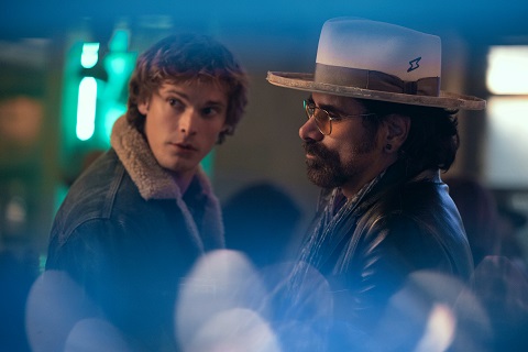 "Two men in a dimly lit indoor setting, one in a jean jacket with a fur collar looking back, the other with a hat and glasses facing forward, colorful lights blurred in the foreground."